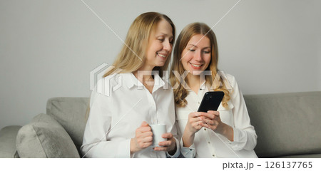 Two women with mobile phone, mature mother and adult daughter together looking at smartphone at home 126137765