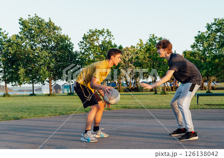 Two Young Caucasian male teenagers Playing Basketball at Park outdoor playground in sunset light. Teenagers' hobby, offline activity. Concept of sports, hobbies and healthy lifestyle. Selective focus. 126138042