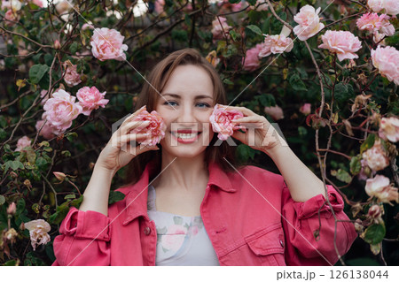 Romantic young woman smiles while holding delicate roses closer to her cheeks amidst beautiful blooming flowers in a vibrant garden. Spring or Summertime vibes, fun, positive mood. Natural skin care 126138044