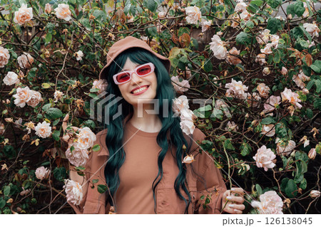 Portrait of young hipster woman in mocha color dress, jacket, bucket hat and sunglasses on blooming roses bush background. Urban street fashion. Gen Z. Trendy fashion outfit. Color of the 2025 year. 126138045