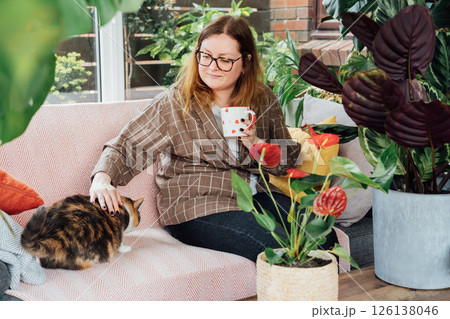 Smiling woman with cup of drink relaxing with cat pet on sofa in home garden conservatory after housekeeping. Portrait of relaxed female resting at home. Gardening hobby. Biophilia lifestyle. 126138046