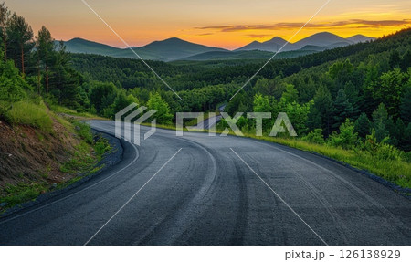 The asphalt road meanders through the forest, leading to distant peaks, while the sunset casts warm orange and pink tones across the sky. 126138929