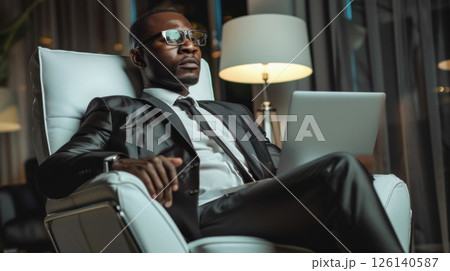 Seated in a stylish modern chair, a man in a tailored suit confidently holds a laptop, embodying the essence of professionalism. 126140587