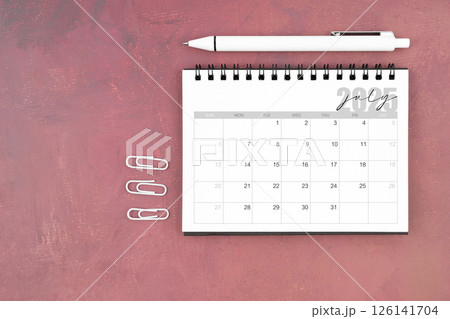 Desk calendar for July 2025 and pen with paper clips on red table. Desk calendar for July 2025 and pen with paper clips on red table. 126141704