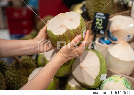 Woman holds green coconut from market stall selecting for milk and dietary use, exotic nut food Woman holds green coconut from market stall selecting for milk and dietary use, exotic nut food 126142611