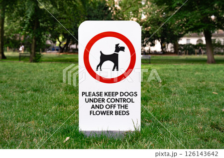 Sign in a park asking to control dogs and keep them off flower beds. Surrounded by green grass trees and public space. 126143642