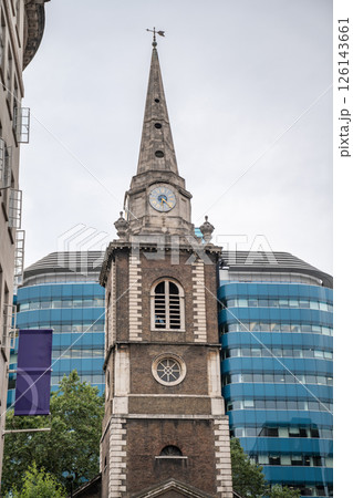 Historic Church Tower with Weathered Stone Spire and Antique Clock Set Against Modern Glass Office Building in Central London, Highlighting Contrast Between Old and New Urban Forms. 126143661