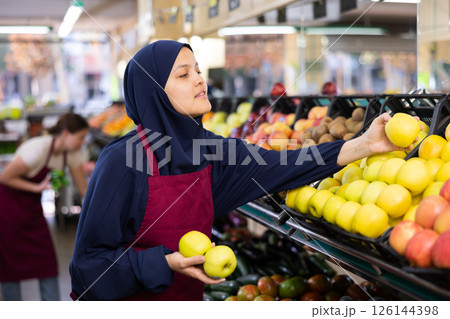 Female seller in hijab and an apron puts fresh apples on shelves in the grocery section of supermarket Female seller in hijab and an apron puts fresh apples on shelves in the grocery section of supermarket 126144398