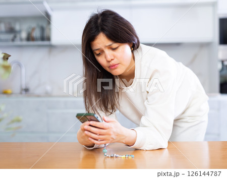 Upset young brunette standing in home kitchen with smartphone 126144787