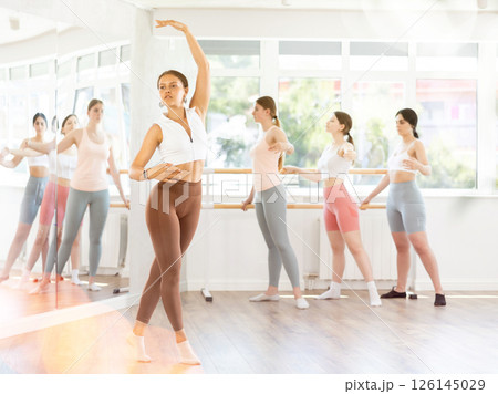 Young beautiful female ballet student demonstrating skills in ballet in dance studio. Group of young ballet dancers in background near ballet barre Young beautiful female ballet student demonstrating skills in ballet in dance studio. Group of young ballet dancers in background near ballet barre 126145029