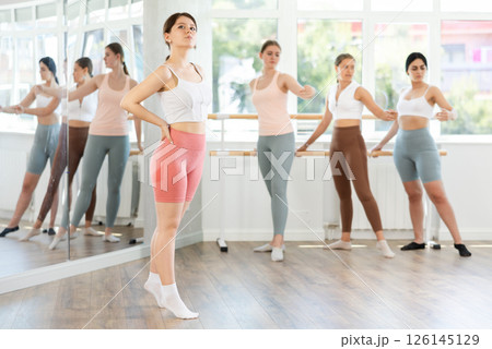 Young beautiful female ballet student demonstrating skills in ballet in dance studio. Group of young ballet dancers in background near ballet barre 126145129