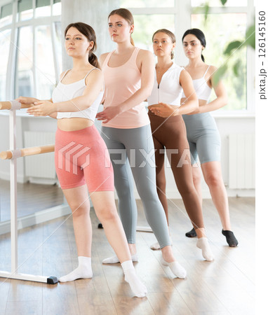 Active young woman practicing battement tendu holding by ballet barre together with others 126145160