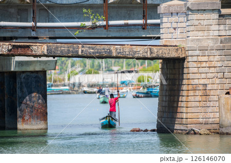 A man in a red shirt rows a small traditional boat under an old, weathered bridge in Mannar, Sri Lanka. Fishing harbor in the background A man in a red shirt rows a small traditional boat under an old, weathered bridge in Mannar, Sri Lanka. Fishing harbor in the background 126146070