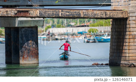 A man in a red shirt rows a small traditional boat under an old, weathered bridge in Mannar, Sri Lanka. Fishing harbor in the background 126146071