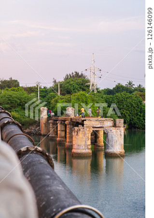 Several people stand on the remains of an old stone bridge over calm water in Mannar, Sri Lanka, surrounded by lush greenery 126146099