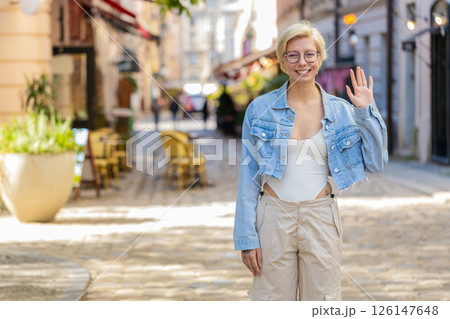 Cheerful young blonde woman smiling friendly at camera waving hands gesturing on city urban street 126147648