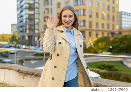 Cheerful mature woman in coat smiling at camera waving hands gesturing hello standing on city street Cheerful mature woman in coat smiling at camera waving hands gesturing hello standing on city street 126147660