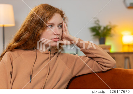 Upset lonely young woman with long red hair thinking about problems sitting on sofa in living room Upset lonely young woman with long red hair thinking about problems sitting on sofa in living room 126147670