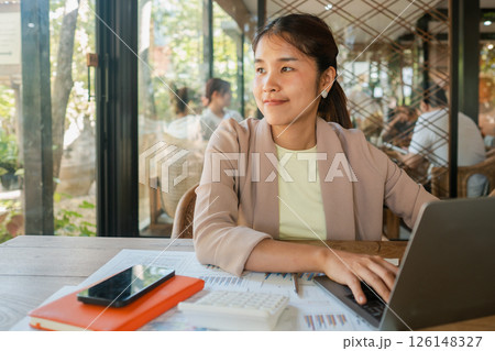 A young woman in a modern cafe, working on a laptop with business documents and a smartphone, surrounded by natural light. A young woman in a modern cafe, working on a laptop with business documents and a smartphone, surrounded by natural light. 126148327