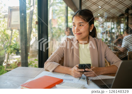 Smiling businesswoman holding a smartphone, sitting at a cafe table with documents and a laptop, enjoying a break. 126148329