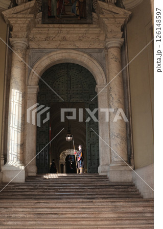 Swiss Guard in traditional Renaissance uniform holding ceremonial halberd under arched marble doorway inside Vatican City with dramatic light and historic architecture guarding entrance to sacred area 126148597