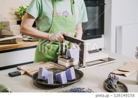 Woman in green apron slicing handmade lavender soap on a worktable with bunches of dried lavender nearby. Clean beauty, natural ingredients, chemical-free soap, small-scale production Woman in green apron slicing handmade lavender soap on a worktable with bunches of dried lavender nearby. Clean beauty, natural ingredients, chemical-free soap, small-scale production 126149158