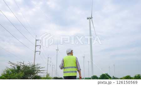 Engineer walking through wind turbine field inspecting renewable energy infrastructure under cloudy sky 126150569