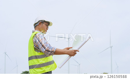 Engineer holding blueprint while surveying wind turbines at renewable energy site under bright sky conditions. 126150588