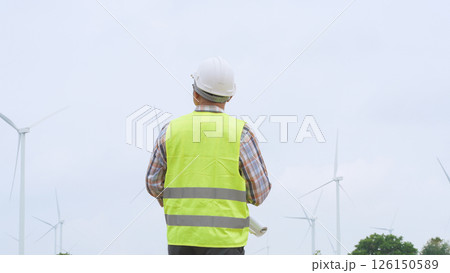Engineer holding blueprint while surveying wind turbines at renewable energy site under bright sky conditions. Engineer holding blueprint while surveying wind turbines at renewable energy site under bright sky conditions. 126150589