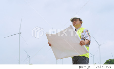 Engineer holding blueprint while surveying wind turbines at renewable energy site under bright sky conditions. 126150595