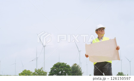 Engineer holding blueprint while surveying wind turbines at renewable energy site under bright sky conditions. 126150597