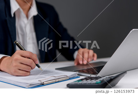 businessman working on papers, taking notes, and analyzing financial data on his laptop. 126153973