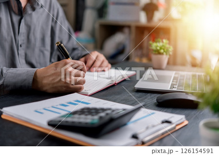 Businessman writing notes while working on financial reports at an office desk with a laptop and calculator. Perfect for business, finance, and productivity concepts. 126154591