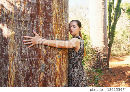 Young woman spreading her arms around huge baobab tree trunk, hugging it, eyes closed 126155474