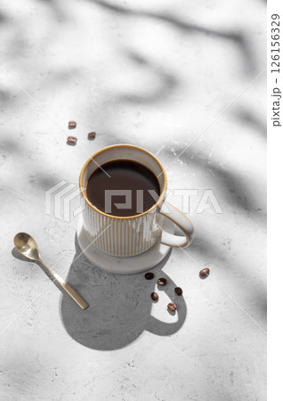 Cup of freshly brewed coffee on light textured background with morning shadows and coffee beans. 126156329