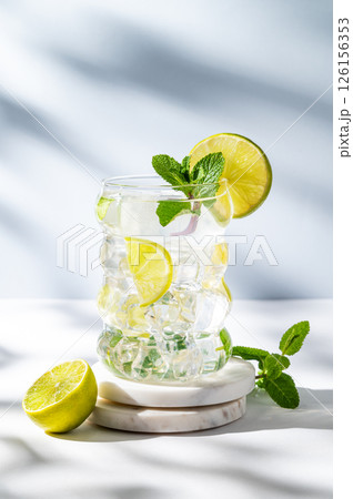 Mojito cocktail in a glass with lime, mint, ice on a marble stand on a light background with shadows Mojito cocktail in a glass with lime, mint, ice on a marble stand on a light background with shadows 126156353