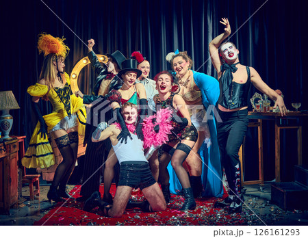 Cabaret cast laughs around central performer with pink boa and hats under spotlight on confetti carpet 126161293