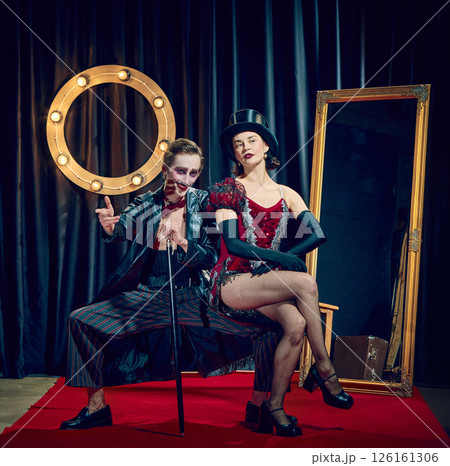 Seated pair of cabaret performers, elegant woman in dress and man with striking makeup, with cane pose against dark retro theatre background. 126161306