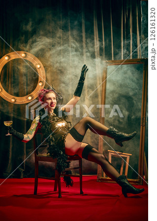 Solo female cabaret performer, eccentric woman in sequined vintage costume sits on chair on red carpet, striking dynamic pose with raised arms and leg. Solo female cabaret performer, eccentric woman in sequined vintage costume sits on chair on red carpet, striking dynamic pose with raised arms and leg. 126161320
