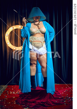Cabaret females performer in distinctive vintage costume featuring lampshade hat and robe stands dramatically on confetti-covered red carpet. Cabaret females performer in distinctive vintage costume featuring lampshade hat and robe stands dramatically on confetti-covered red carpet. 126161329