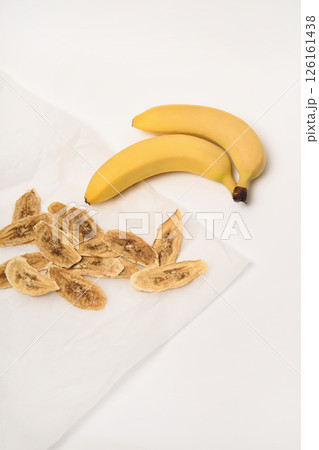 Two fresh bananas, next to which are dried banana slices on parchment paper. White background. 126161438