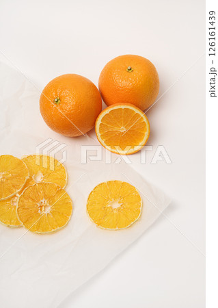 Two fresh oranges, next to which are dried orange slices on parchment paper. White background. 126161439