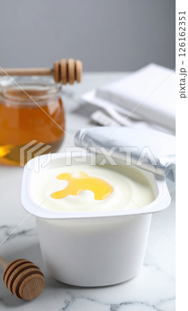 Creamy yogurt with honey drizzle in white container, accompanied by honey jar and dipper on marble surface 126162351