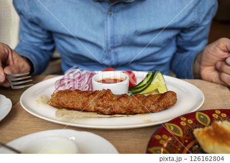 Grilled kebab served with fresh vegetables and dipping sauce on a white plate in a dining setting Grilled kebab served with fresh vegetables and dipping sauce on a white plate in a dining setting 126162804