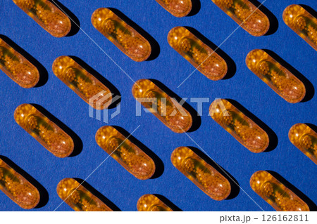 Pattern of yellow transparent liquid capsules with granules on a blue background. Medicaments aesthetically pleasing for advertising. Pattern of yellow transparent liquid capsules with granules on a blue background. Medicaments aesthetically pleasing for advertising. 126162811