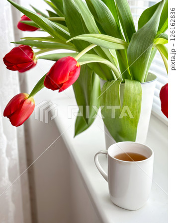 A cozy morning scene with a white ceramic cup of tea placed on a windowsill next to a bouquet of red tulips in a white vase 126162836
