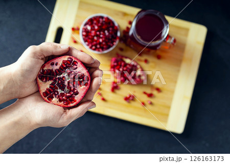 Hands holding cutted pomegranate and a ceramic bowl full of pomegranate seeds, cutted pomegranate and a glass of pomegranate juice on the background lie on the wooden cutting board. View from the top 126163173