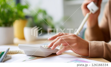 Person communicating on a landline phone at a desk filled with plants and office supplies during daylight hours 126166334