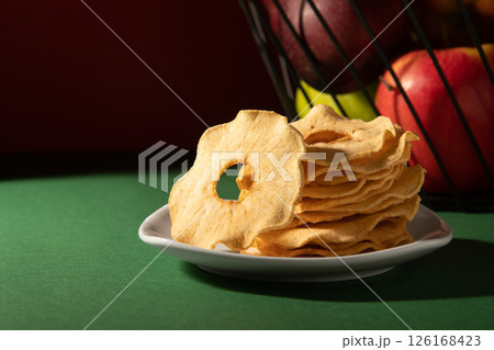 Dried apples on a white plate. Behind is a basket of apples. Studio photo on a dark background. 126168423