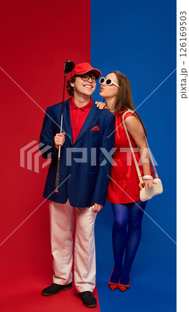 Woman blowing kiss to man smiling holding golf club on red blue split background wearing colorful outfits. 126169503